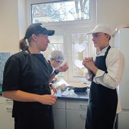 Uczniowie w fartuchach i czapkach rozmawiają ze sobą w pracowni kucharskiej.