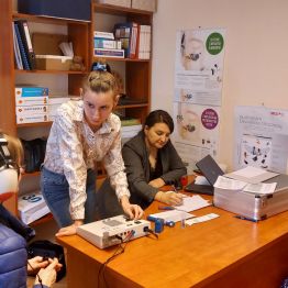 Kobieta w słuchawkach w trakcie badania słuchu, obok dwie protetyczki. 