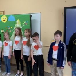 Uczniowie z klas I-III SP stoją ustawieni w rzędzie, recytują wierszyki z okazji Dnia Mamy i Taty.