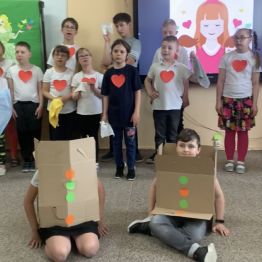 Uczniowie z klas I-III SP inscenizują piosenkę "Mama i Tata to nie są roboty". W centralnej części zdjęcia stoi dwoje dzieci przebranych za roboty, pozostali uczniowie stoją w półkolu.