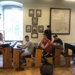 Julia w roli nauczycielki siedzi za biurkiem. Pozostali uczniowie siedzą w ławkach.