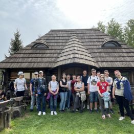 Grupa uczniów przed drewnianą chatą.