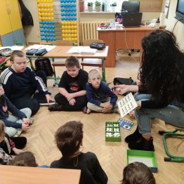 Nauczyciel siedzący z grupą uczniów klasy 1 a i 1 c , wyjaśniający instrukcję kodowania drogi dla ozobota.