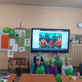 Uczniowie trzymający zielone balony kucają pod ekranem, na którym wyświetlone są osoby przebrane za skrzaty.