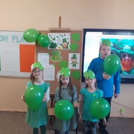 Grupka uczniów w zielonych ubraniach trzyma w rękach zielone balony. W tle flaga Irlandii.