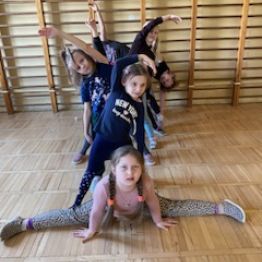 Dziewczynka wykonuje szpagat a pozostałe dzieci inne figury gimnastyczne.