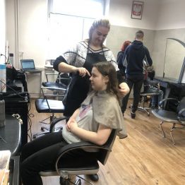 Magda z kl. 3b BS stylizuje fryzurę Oli z kl. 3b BS. Ola siedzi na fotelu fryzjerskim. Z tyłu Kacper i Ola czeszą Krzysia.