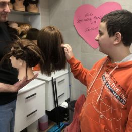 Pan Paweł pokazuje Wojtkowi peruki. Wojtek dotyka prawą ręką jedną z fryzur.