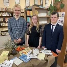 Troje uczniów w szkolnej bibliotece. Przed nimi na stoliku leżą tomiki poezji Wisławy Szymborskiej. 