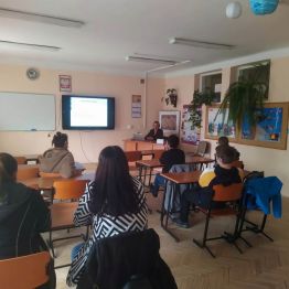 rodzice siedzą w ławkach, w tle tablica interaktywna, biurko i nauczyciel