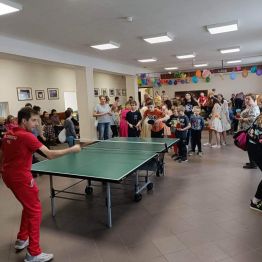 Sportowiec i uczeń grają w tenisa stołowego. Za nimi inni uczniowie przyglądają się rozgrywce.