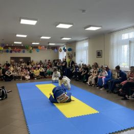 Sportowcy judo w trakcie wykonywania przewrotów na macie. Za nimi widownia.