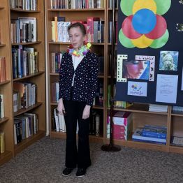 Uczennica recytująca wiersz w bibliotece szkolnej. W tle regał z książkami. 