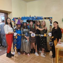 Grupa uczniów w kapeluszach wróżek i wróży wraz z wychowawczyniami pozuje do zdjęcia w „fotobudce”. 