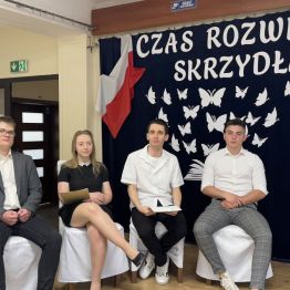 Absolwenci siedzą na scenie. W tle napis: Czas rozwinąć skrzydła.
