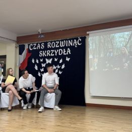 Absolwenci siedzą na scenie i oglądają prezentację zdjęć.