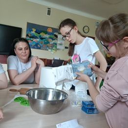 Emilka wsypuje mąkę do miski. Karolina z panią studentką obserwują.
