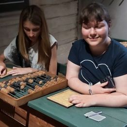 dziewczynki siedzą w ławkach szkolnych na których są liczydła i dawny zeszyt do pisania