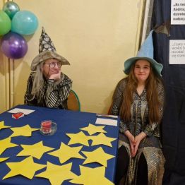 Wróżbici siedzą przy stanowisku: „losowanie gwiazdek” oraz przebicie balona z przepowiednią.