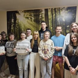 Wychowawca i grupa wychowanków stojących razem w pomieszczeniu na tle leśnej grafiki, z których jedna trzyma kartkę z napisem „SMUTEK”.