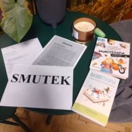 Na zdjęciu widać stolik z książkami o bajkoterapii, kartką z napisem „SMUTEK”, świecą i rośliną, tworzące spokojną, refleksyjną atmosferę.