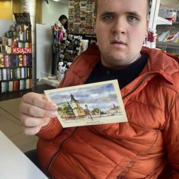 Kamil siedzi. W ręce trzyma widokówkę z widokiem naszego miasta.
