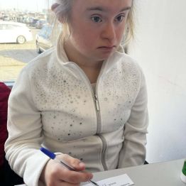 Kasia siedzi. W ręce trzyma długopis. Przed nią leży wypisana kartka z życzeniami. 