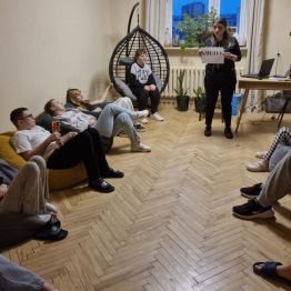 Wychowawca przedstawia temat dzisiejszego spotkania, trzyma w ręku kartkę z napisem „SMUTEK”, wychowankowie leżą w kręgu na pufach.