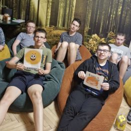 Wychowawca i grupa wychowanków siedzi wygodnie na pufach, w pomieszczeniu na tle leśnej grafiki, z których dwóch wychowanków trzyma kartkę graficzną związaną z radością.