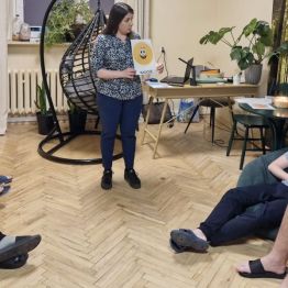 Wychowawca przedstawia temat dzisiejszego spotkania, trzyma w ręku kartkę z napisem „RADOŚĆ”, wychowankowie leżą w kręgu na pufach.