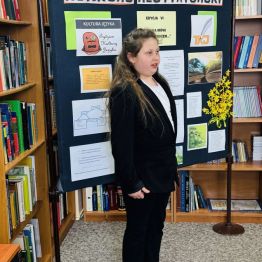 Uczennica recytująca wiersz. W tle tablica informacyjna.