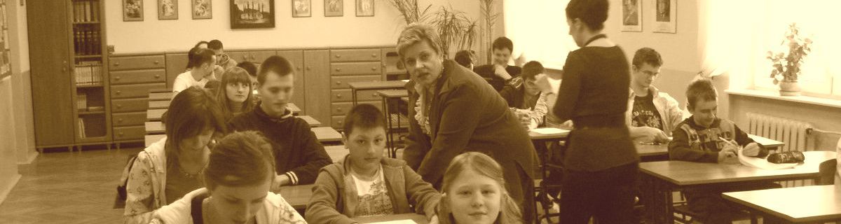 Uczniowie siedzą w ławkach, obok jednej osoby stoi nauczycielka i coś tłumaczy
