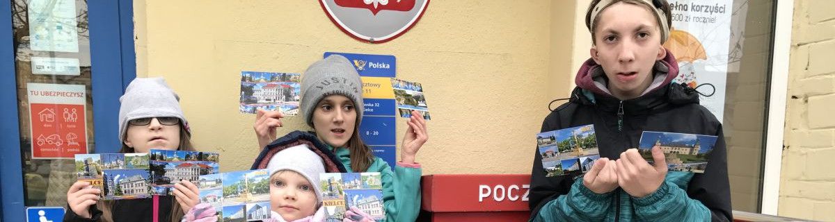 Uczniowie stoją przed pocztą i trzymają kartki pocztowe