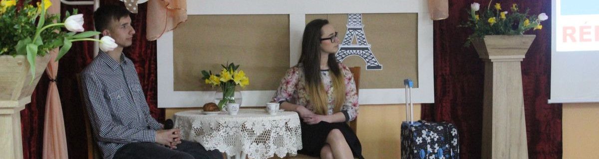 Uczeń i uczennica siedzą na scenie przy stoliku nakrytym koronkowym obrusem, obok stoi walizka.