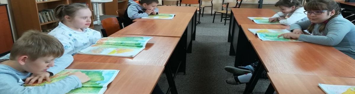Uczniowie siedzą w ławkach i przeglądają atlasy geograficzne
