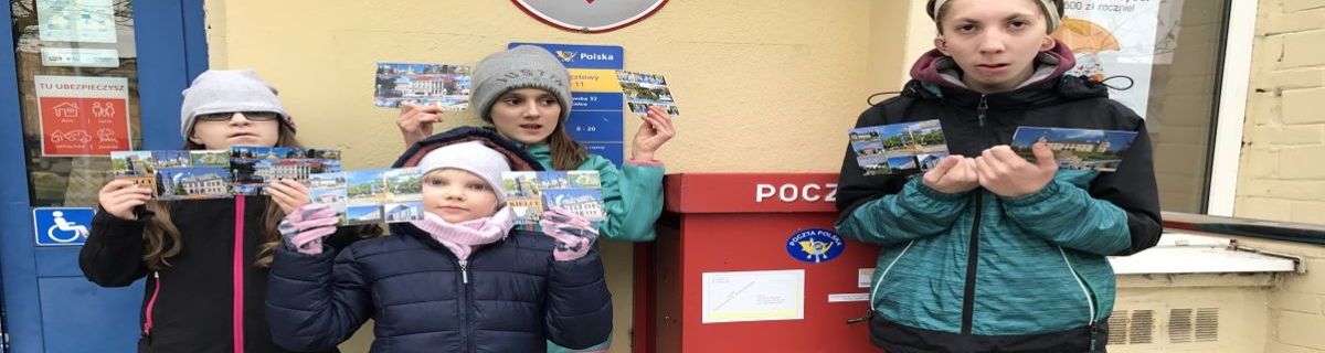 Uczniowie stoją przed pocztą i trzymają w rękach kartki pocztowe