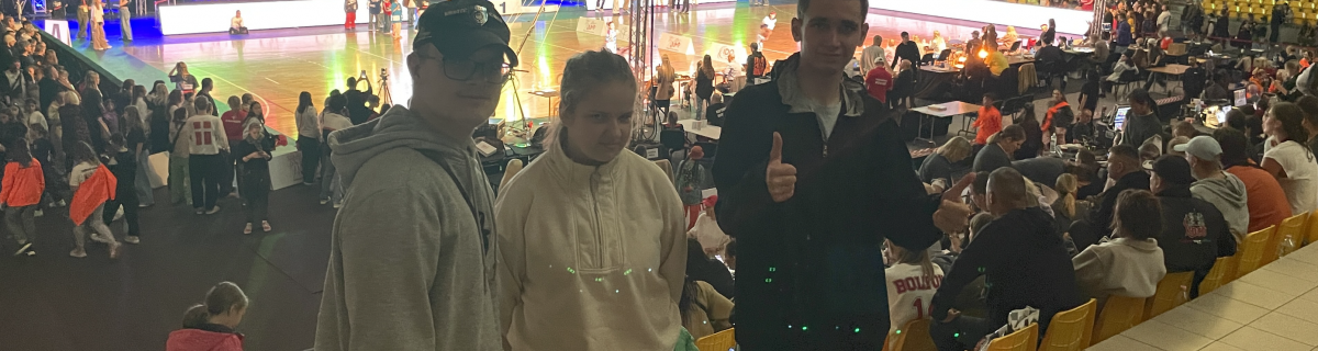 Uczniowie na widowisku tanecznym w hali sportowej. W tle płyta boiska i tancerze.