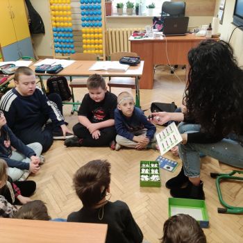 Nauczyciel siedzący z grupą uczniów klasy 1 a i 1 c , wyjaśniający instrukcję kodowania drogi dla ozobota.