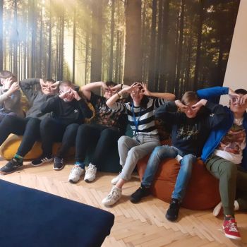 Uczniowie i wychowawczyni siedzą na pufach w sali terapeutycznej. W tle fototapeta przedstawiająca las.