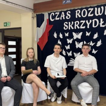 Absolwenci siedzą na scenie. W tle napis: Czas rozwinąć skrzydła.