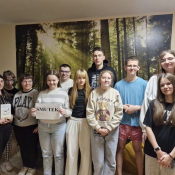 Wychowawca i grupa wychowanków stojących razem w pomieszczeniu na tle leśnej grafiki, z których jedna trzyma kartkę z napisem „SMUTEK”.
