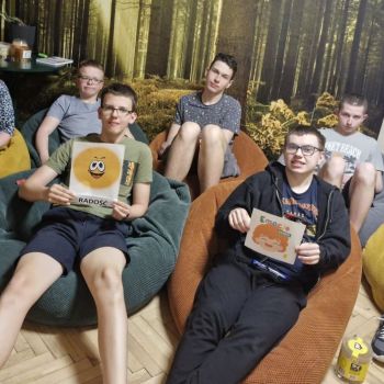 Wychowawca i grupa wychowanków siedzi wygodnie na pufach, w pomieszczeniu na tle leśnej grafiki, z których dwóch wychowanków trzyma kartkę graficzną związaną z radością.