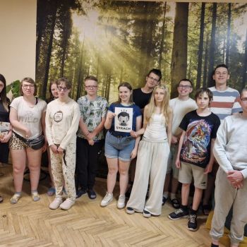 Wychowawca i grupa wychowanków stoi w pomieszczeniu na tle leśnej grafiki, jedna z dziewczynek trzyma kartkę graficzną związaną z emocją strachu.